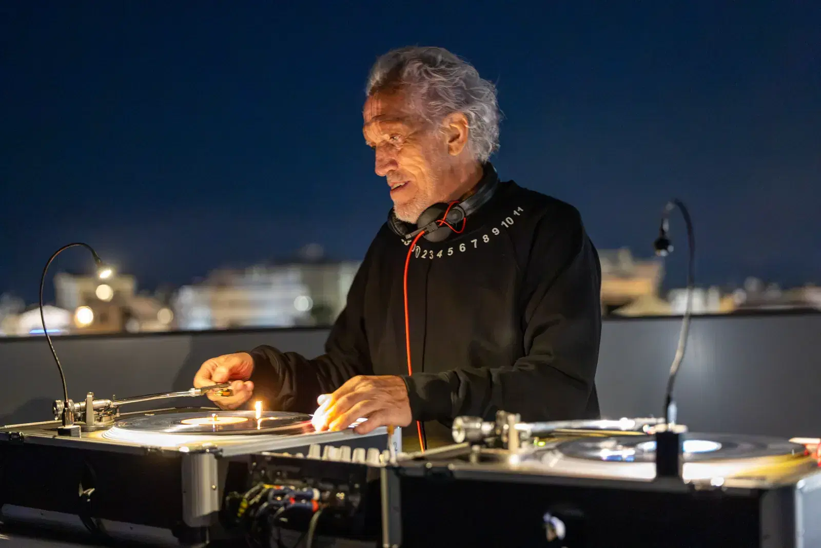 A man with headphones around his neck at the DJ console with turntables, on the terrace in the evening, with the city lights behind