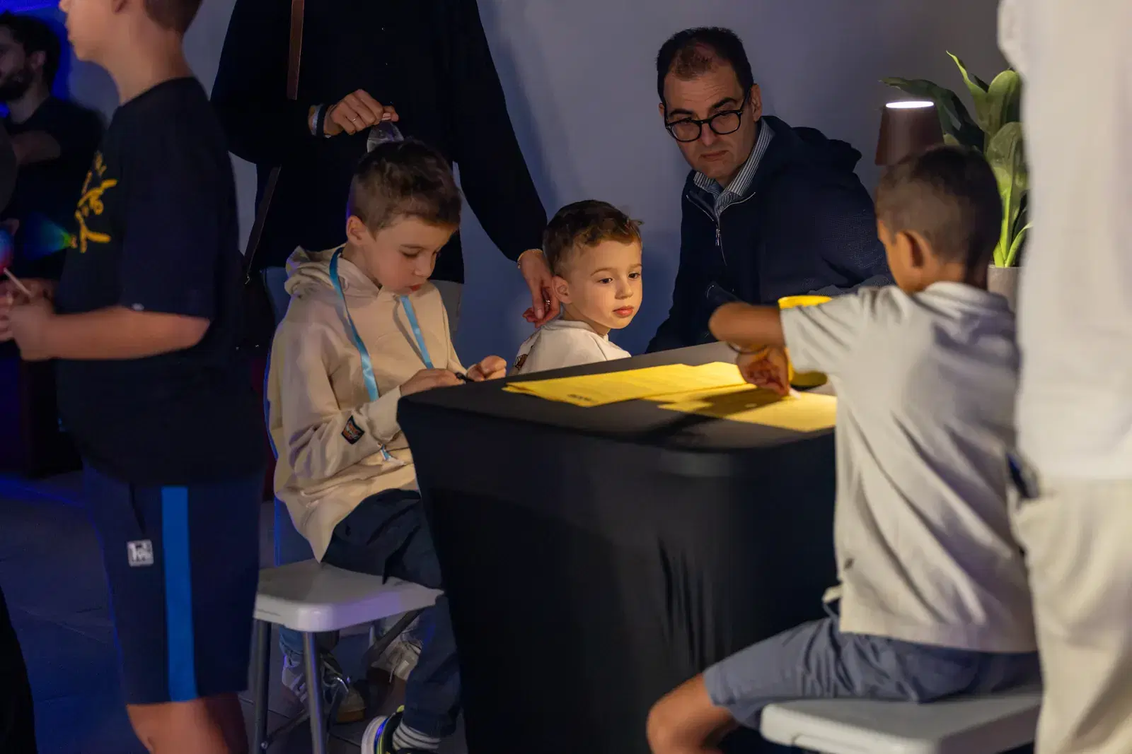 Three children draw at a table, with an adult sitting nearby and more children around