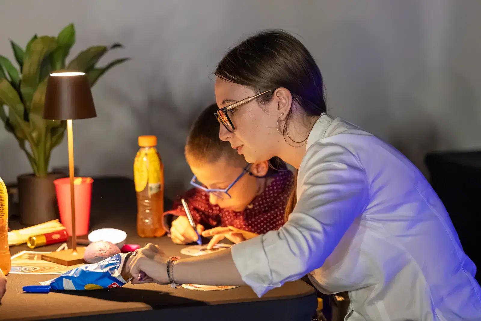 A young woman in a white shirt helps a boy with glasses draw, at a table lit by a lamp