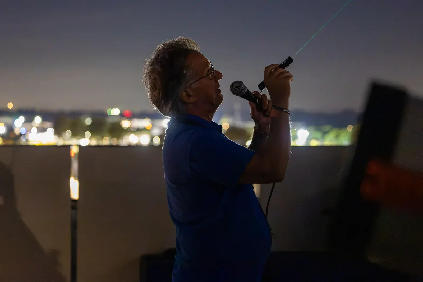 A man speaks into a microphone and points a green laser at the sky from the terrace, with the lights of Jesolo behind him