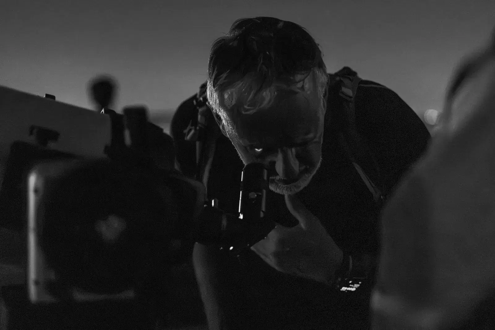 In black and white, a man bends over the eyepiece of a telescope in the dark of the terrace