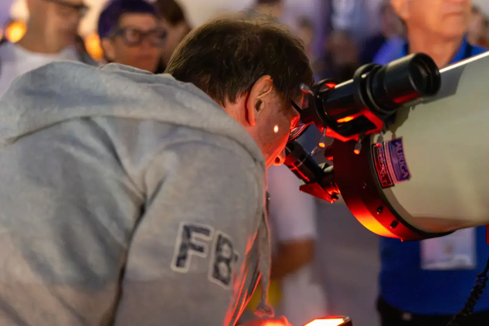 A man in a grey hoodie bends over the eyepiece of a white telescope, lit by red light, with other people waiting