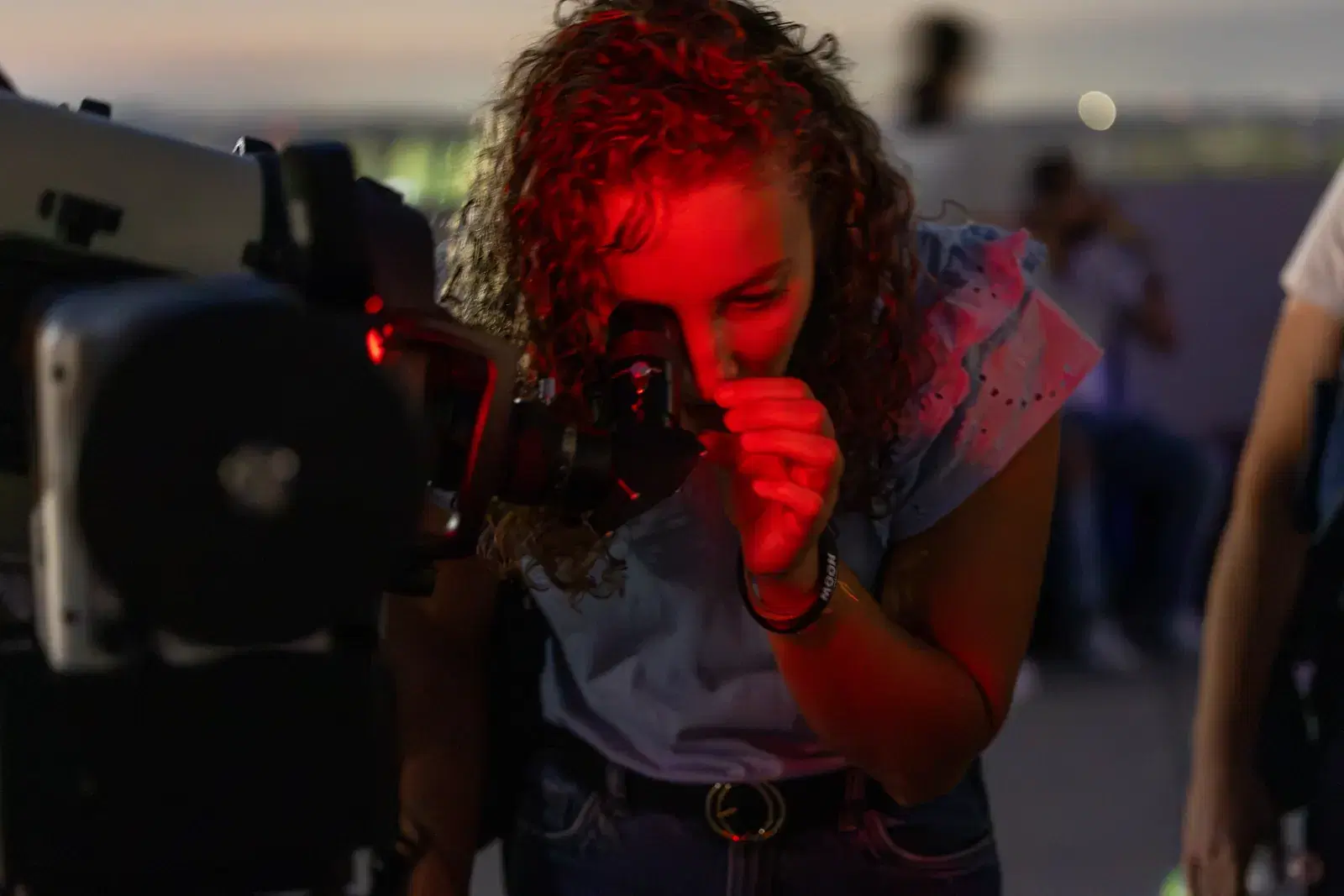 A curly-haired girl, lit by red light, looks into the eyepiece of a telescope