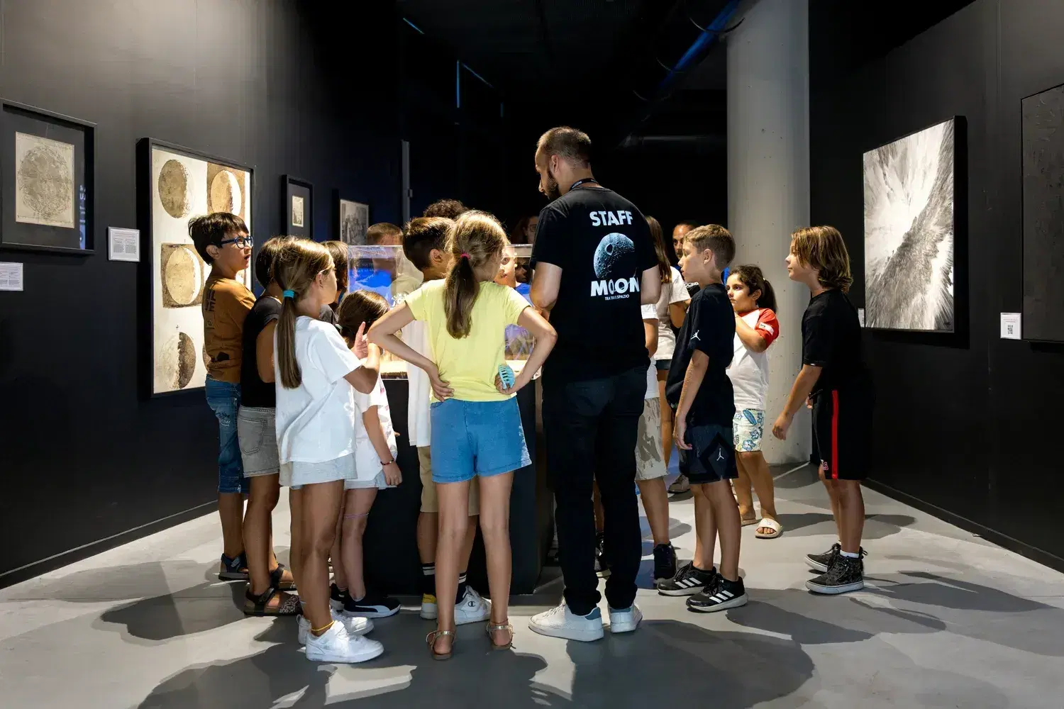 A group of visitors, adults and children, listens to a guide in a MOON T-shirt in one of the exhibition rooms
