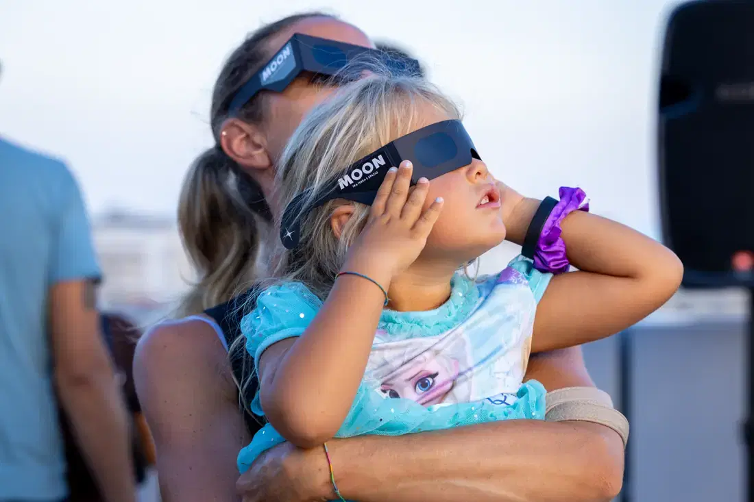 Una bambina in braccio guarda il Sole con gli occhiali da eclissi, accanto a lei una donna con gli stessi occhiali