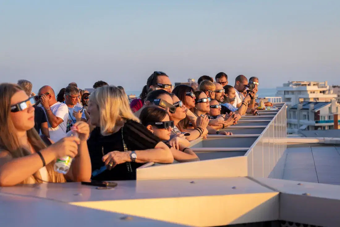 Una lunga fila di persone con gli occhiali da eclissi appoggiate al parapetto della terrazza