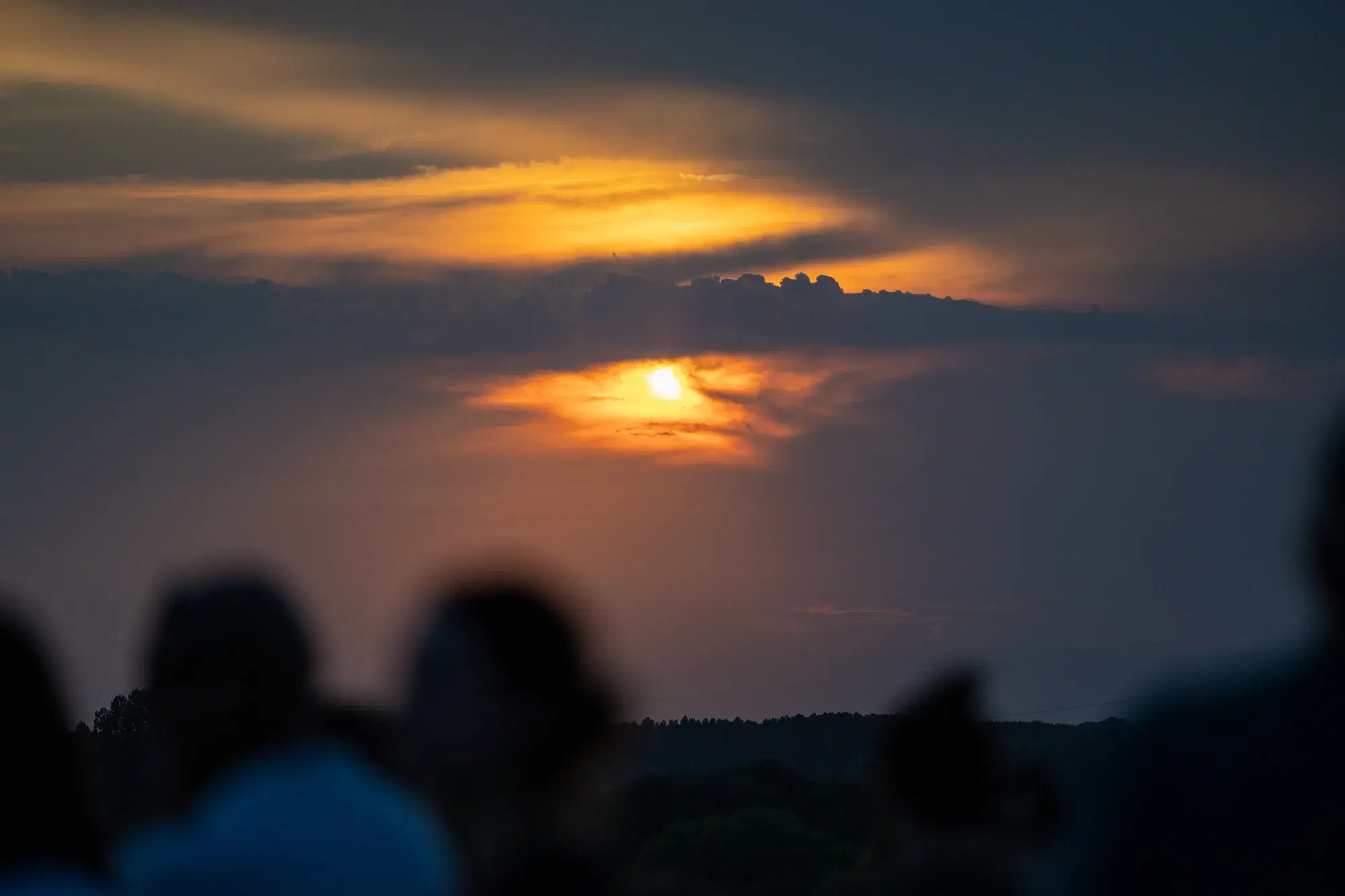 Il Sole basso all'orizzonte filtra tra le nuvole arancioni sopra le sagome del pubblico