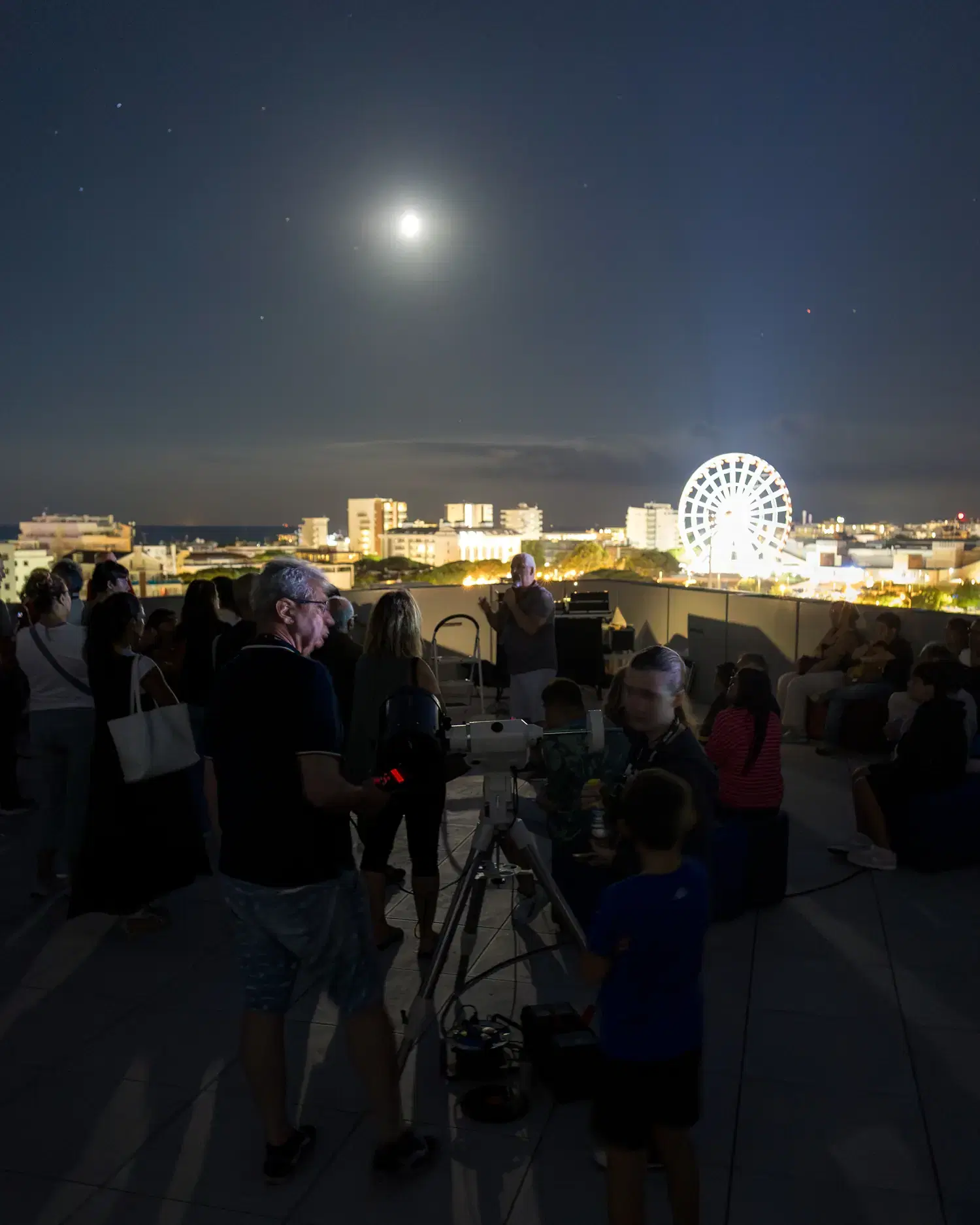 La Luna alta nel cielo notturno sopra la ruota panoramica di Jesolo, con il pubblico e un telescopio sulla terrazza