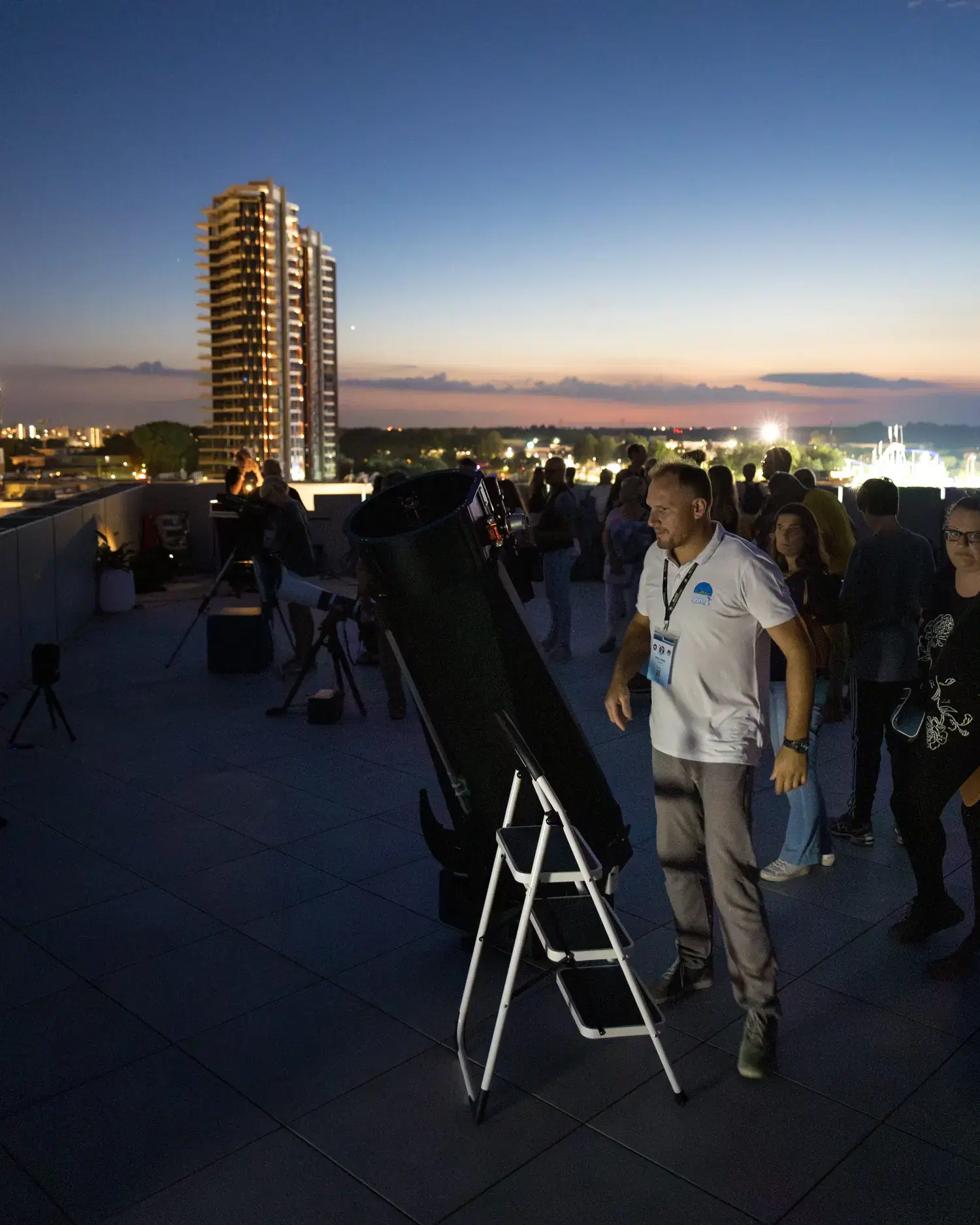 Un telescopio newtoniano con la scaletta accanto sulla terrazza, davanti al grattacielo di Jesolo illuminato nell'ora blu