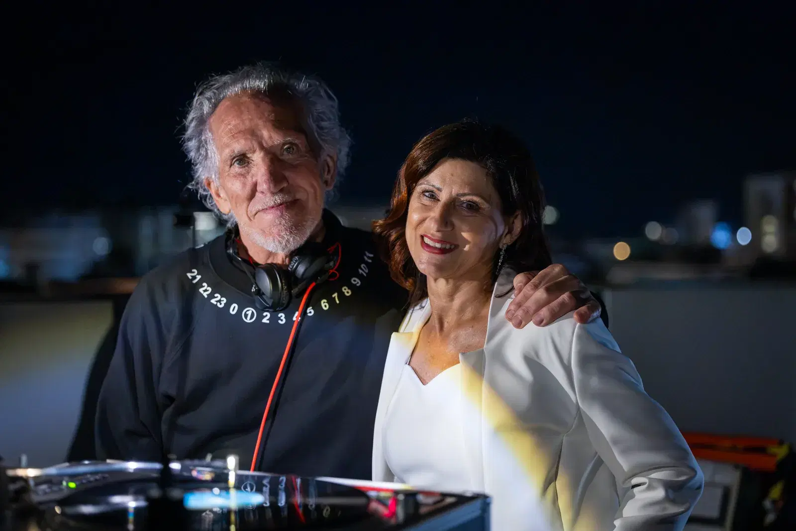 Due persone dietro la consolle del dj sulla terrazza, di sera, con le luci della città alle spalle