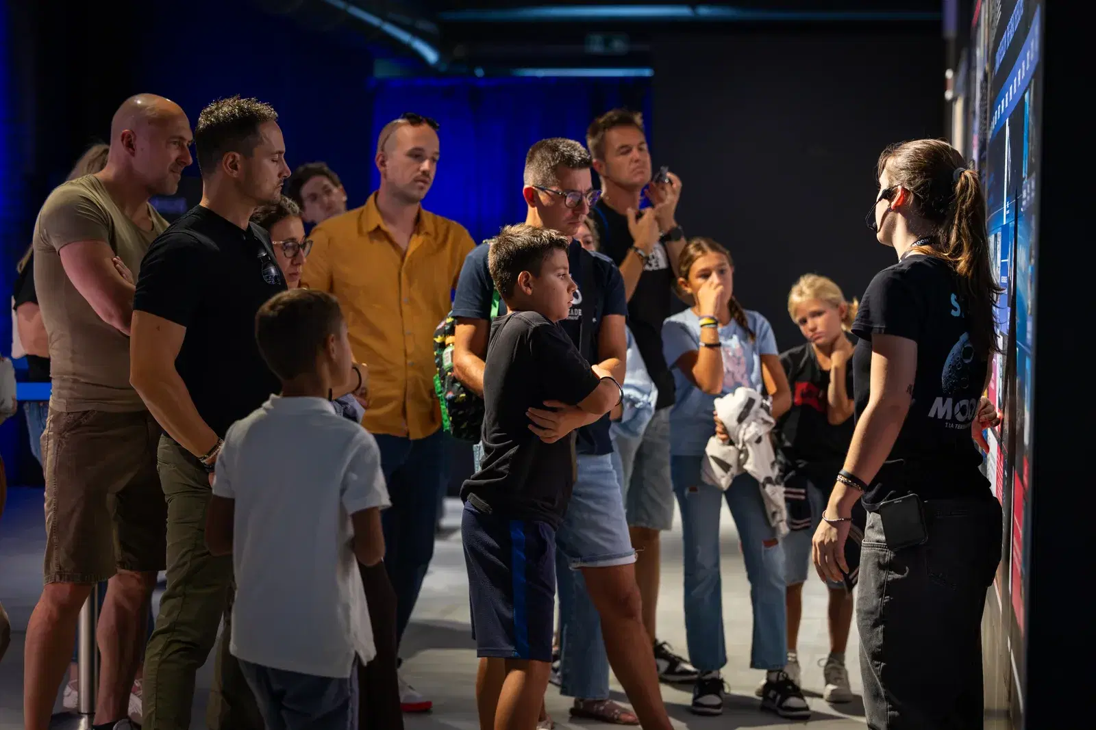 Un gruppo numeroso di visitatori, adulti e bambini, ascolta una guida in maglietta MOON in una sala buia