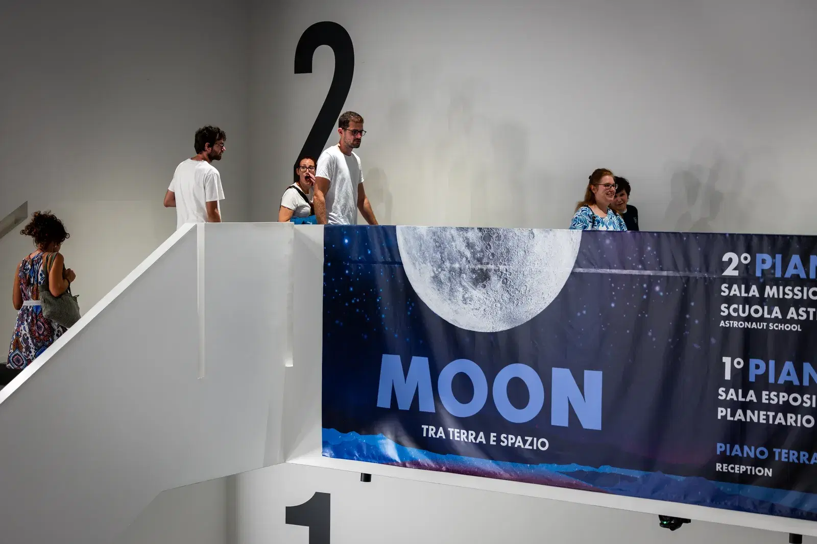L'interno del JMuseo con il cartello MOON — Tra Terra e Spazio e l'indicazione dei piani, e i visitatori sul ballatoio