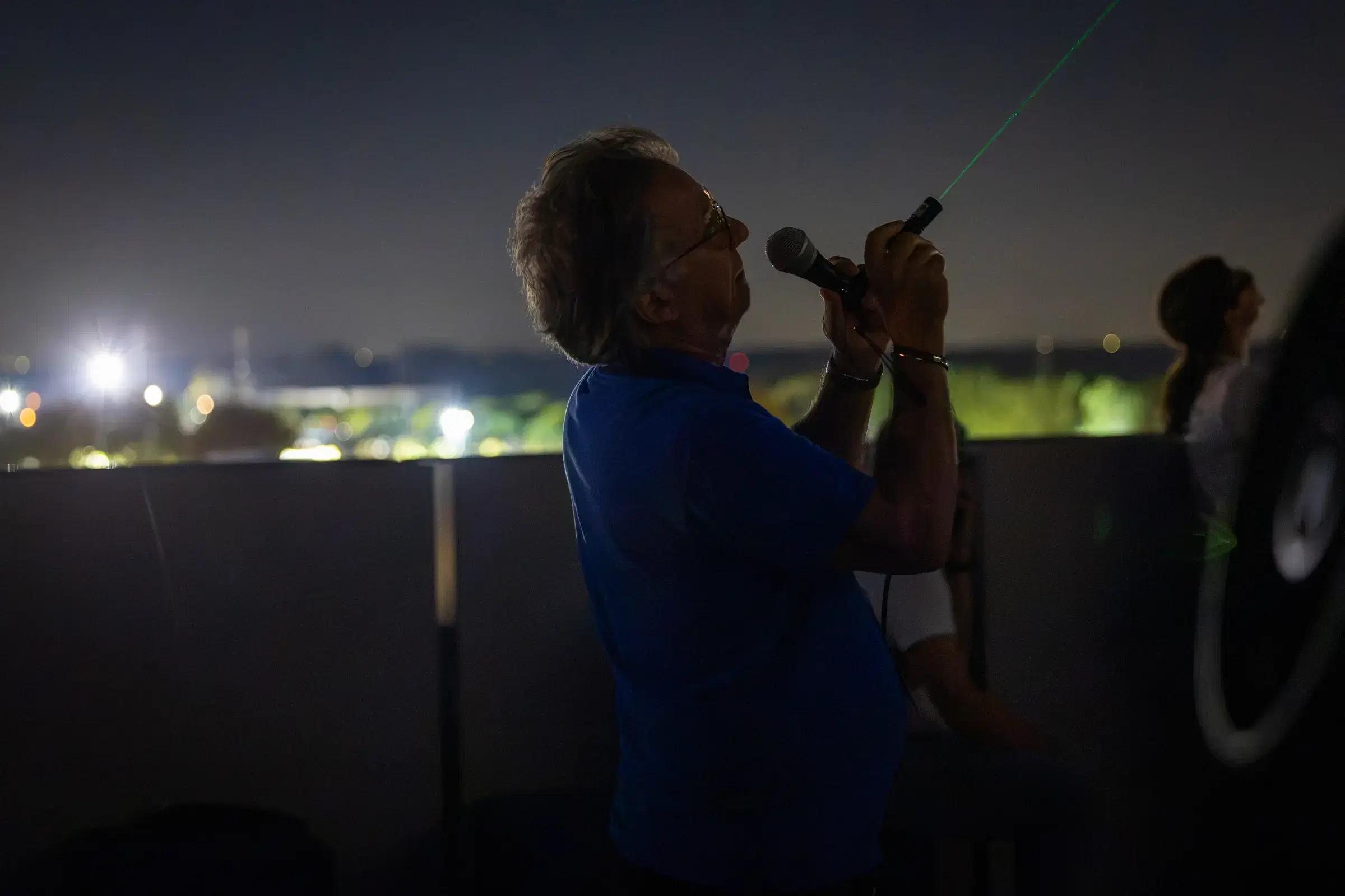 Un astrofilo parla al microfono e indica il cielo con un puntatore laser verde dalla terrazza del JMuseo, con le luci di Jesolo alle spalle