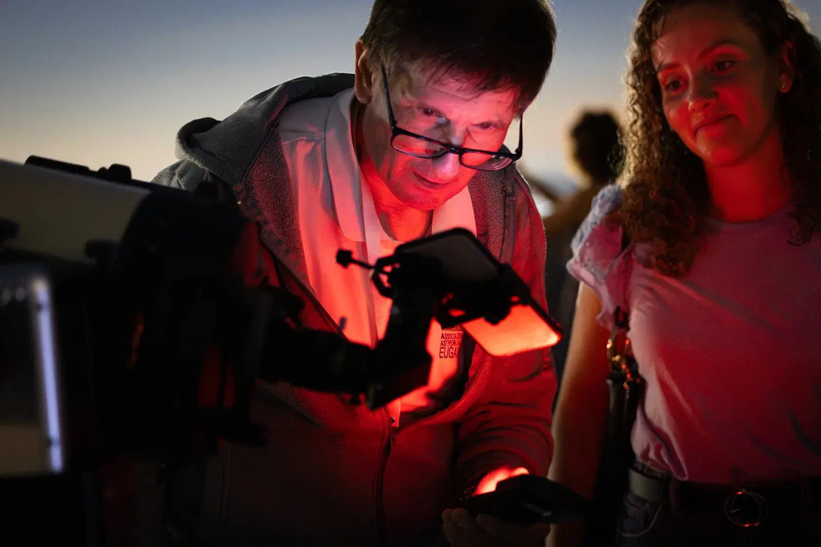 Alla luce rossa di una torcia, un visitatore all'oculare del telescopio e una ragazza accanto, con il cielo del tramonto alle spalle