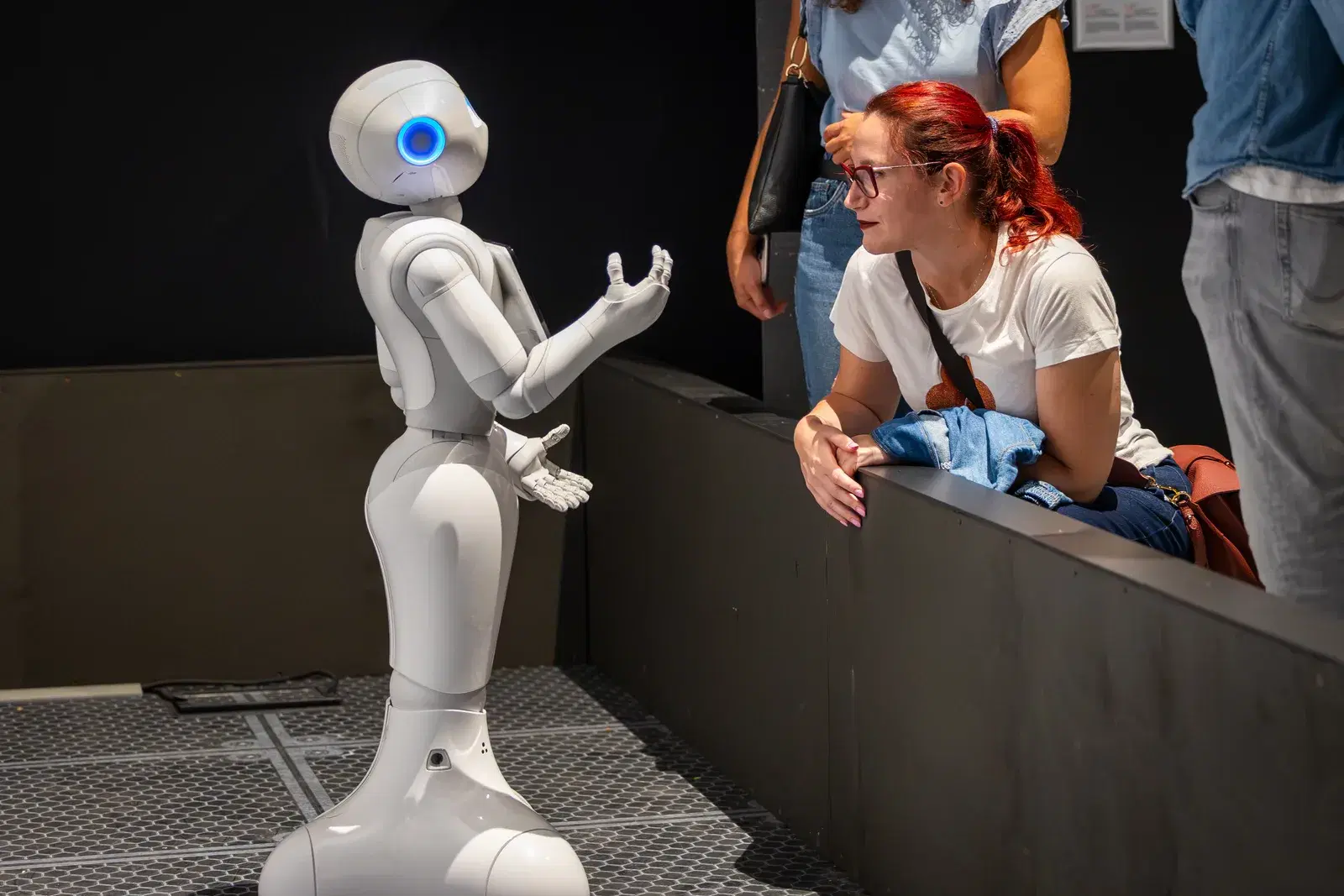Il robot umanoide bianco della mostra gesticola davanti a una visitatrice che lo ascolta appoggiata al parapetto