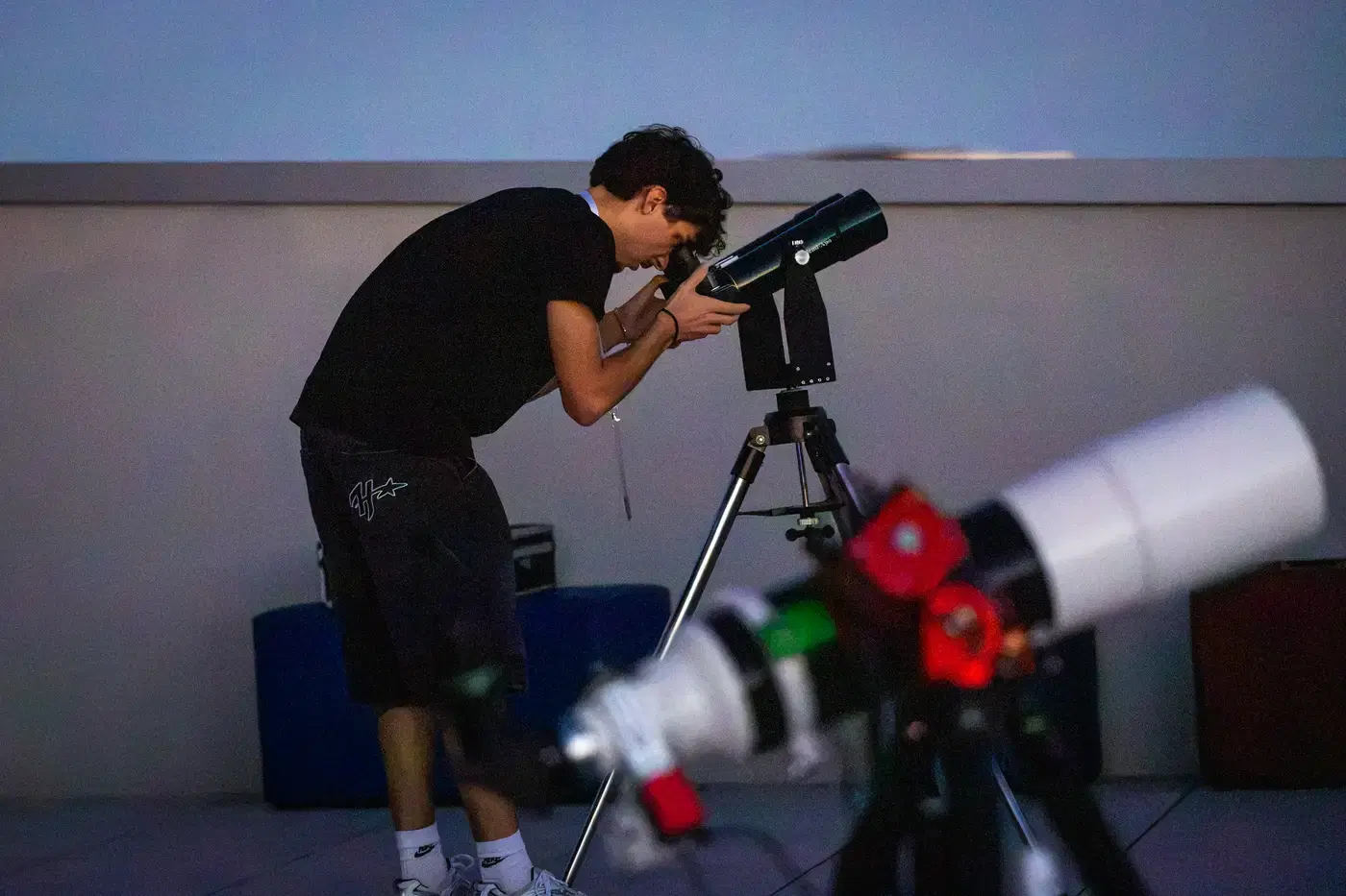 Un ragazzo punta il telescopio chinandosi sullo strumento, in terrazza, con la luce del giorno ancora alta